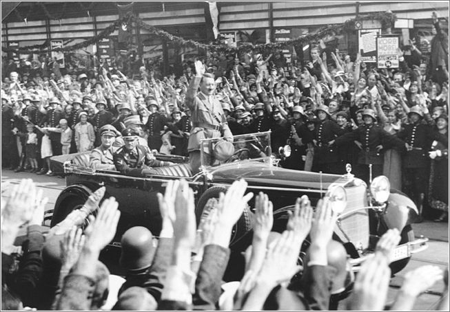 Standing in an open car, Adolf Hitler salutes a crowd lining the streets of Hamburg